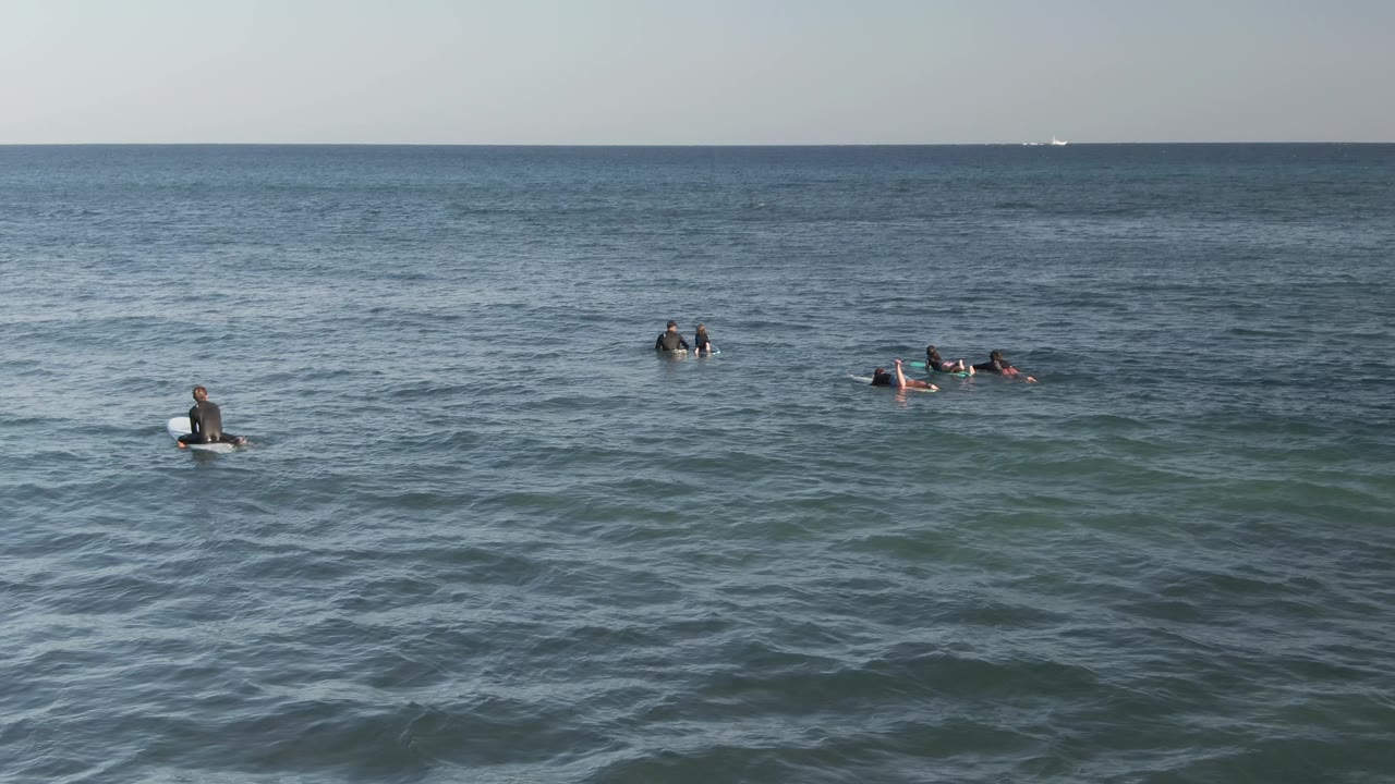 Wide elevated view of surfers spread across calm ocean water at Costa Azul Los Cabos, Mexico, with low chop, pale sky, and distant vessels on the horizon.