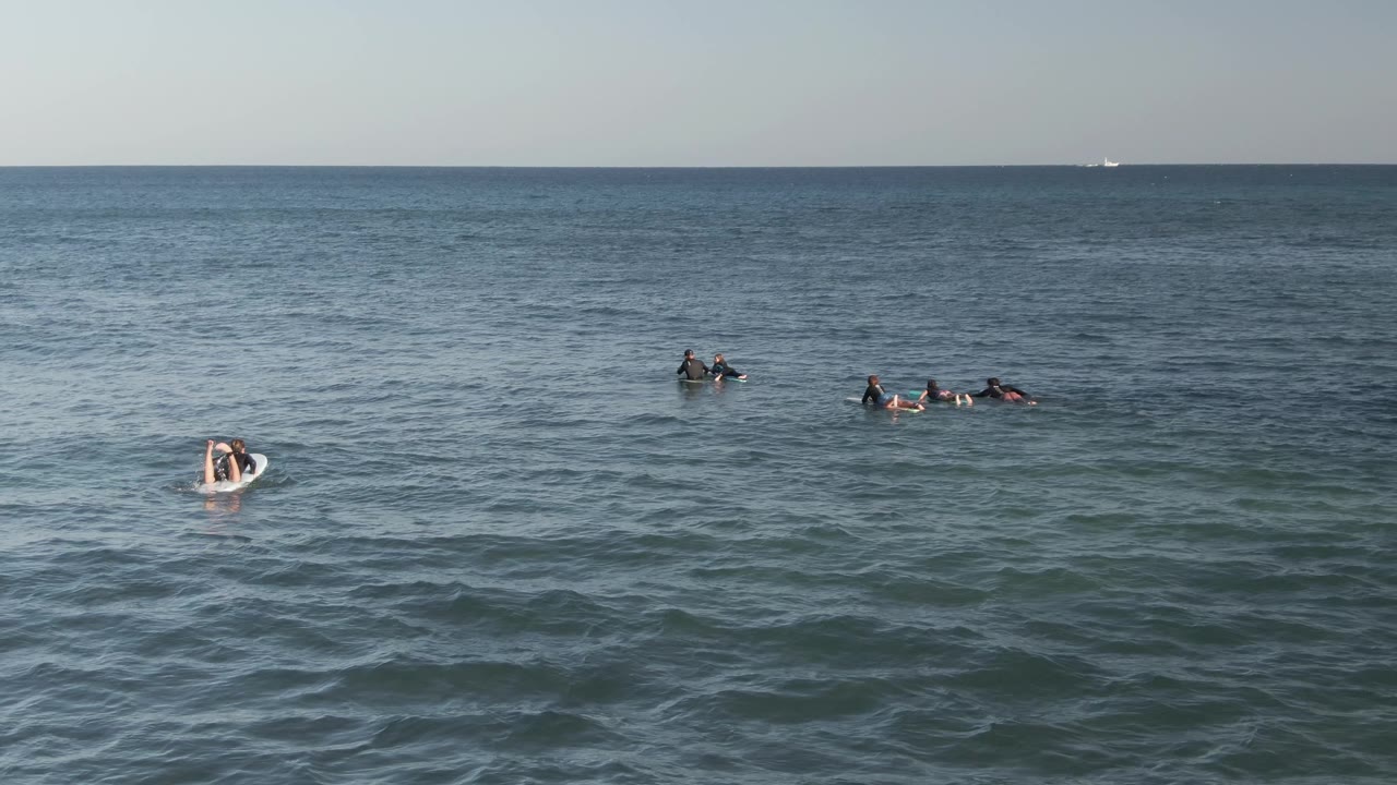 Several small surfer groups remain offshore at Costa Azul Los Cabos, Mexico, while gentle waves texture the foreground and the horizon line stays level in the distance.