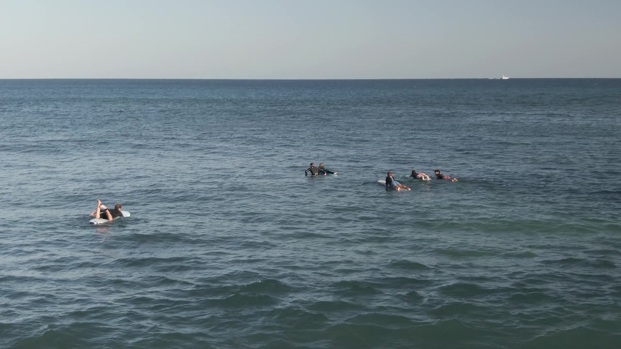 Surfers on boards are spread across open water with a wide foreground of waves.