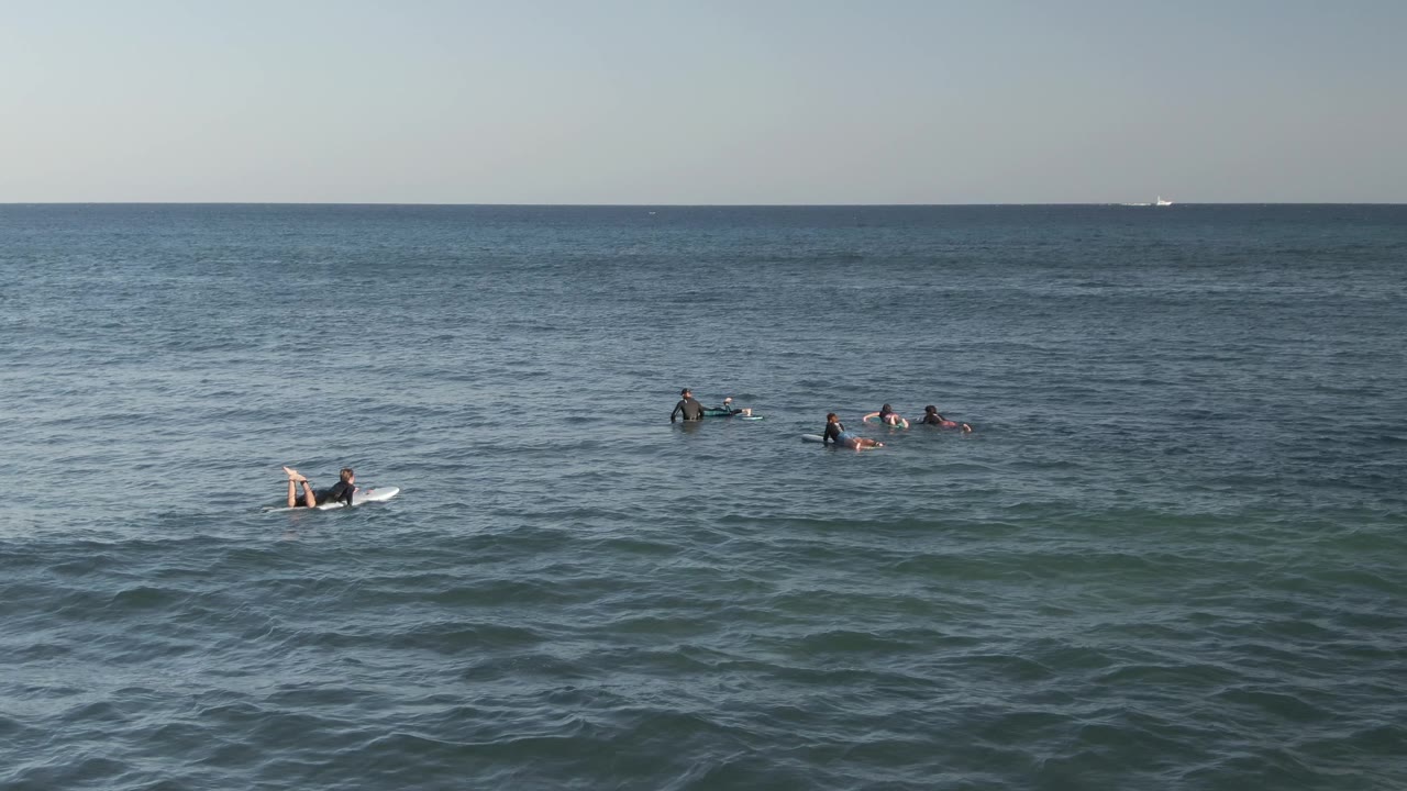 Wide view of surfers spread across calm blue-green ocean water at Costa Azul Los Cabos, with a pale sky and distant boats on the horizon.
