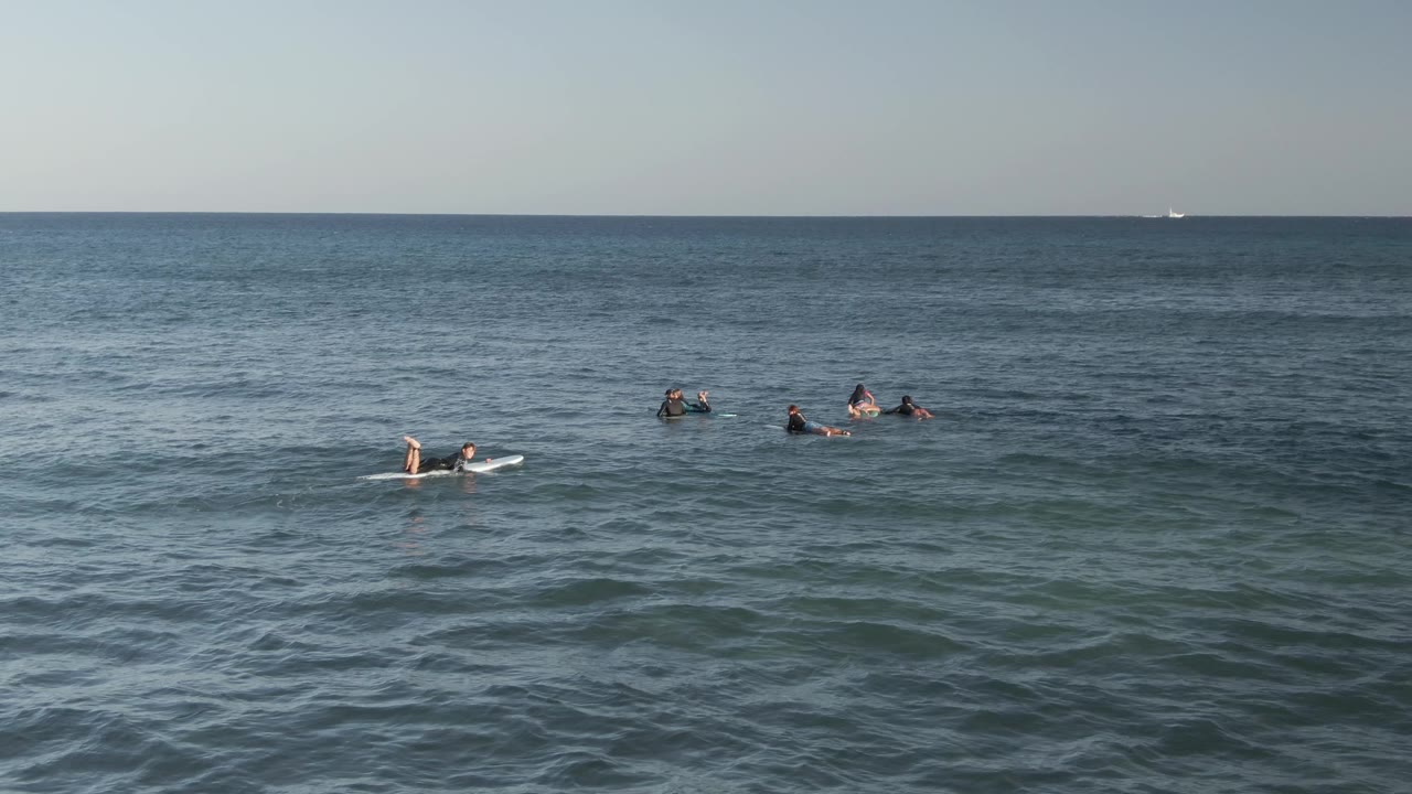 A wide ocean frame at Costa Azul Los Cabos shows several surfers clustered and separated on boards, with gentle texture on the water and faint boats near the horizon.