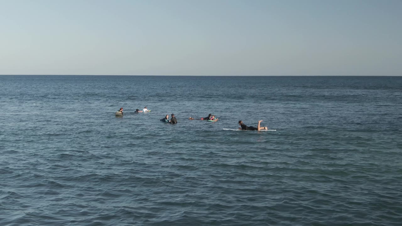 At Costa Azul Los Cabos, surfers and bodyboarders are spaced across the midground of a wide ocean view with a clear horizon and rippled blue water.
