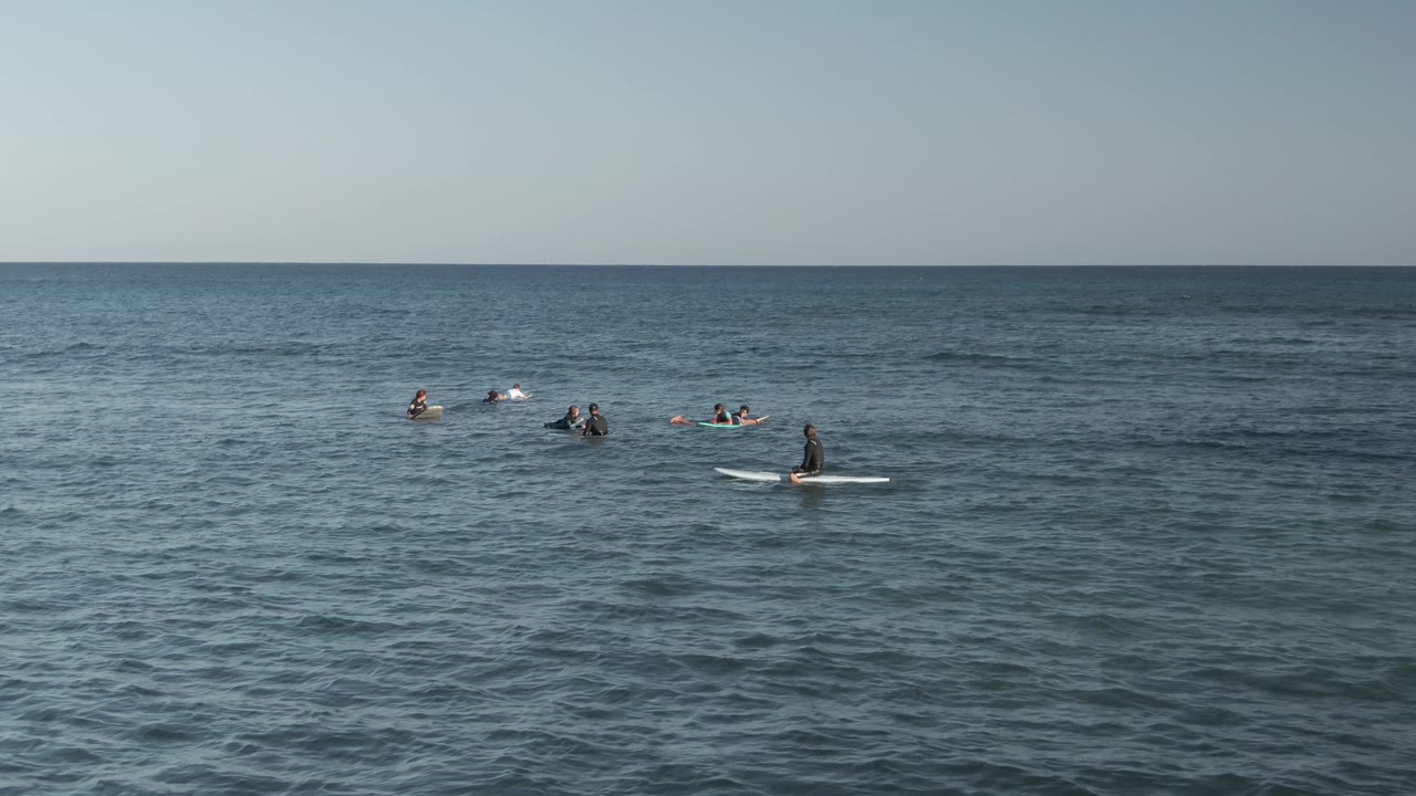 At Costa Azul Los Cabos, the surf lineup is spread across the upper half of the frame, with a seated surfer on a white board to the right and textured water below.