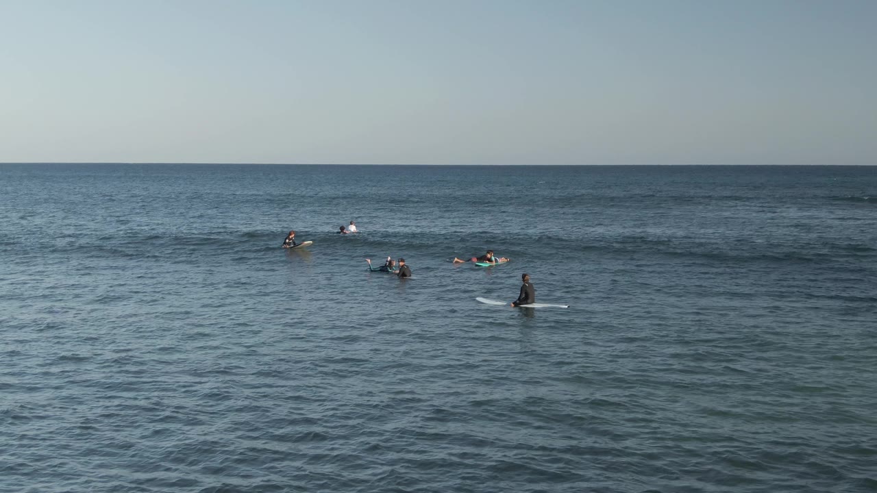 Wide view of surfers on surfboards waiting in the ocean at Costa Azul Los Cabos, with blue water in the foreground and a clear horizon above them.