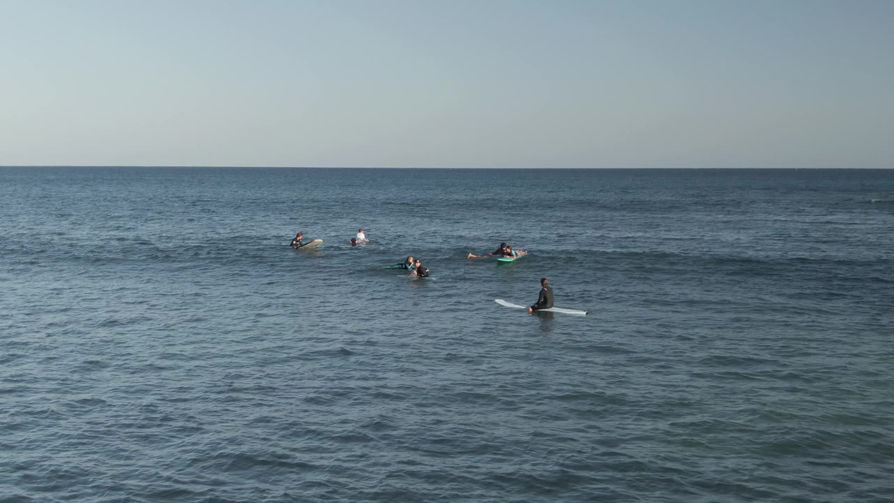 Several surfers sit and paddle on surfboards beyond a small swell.