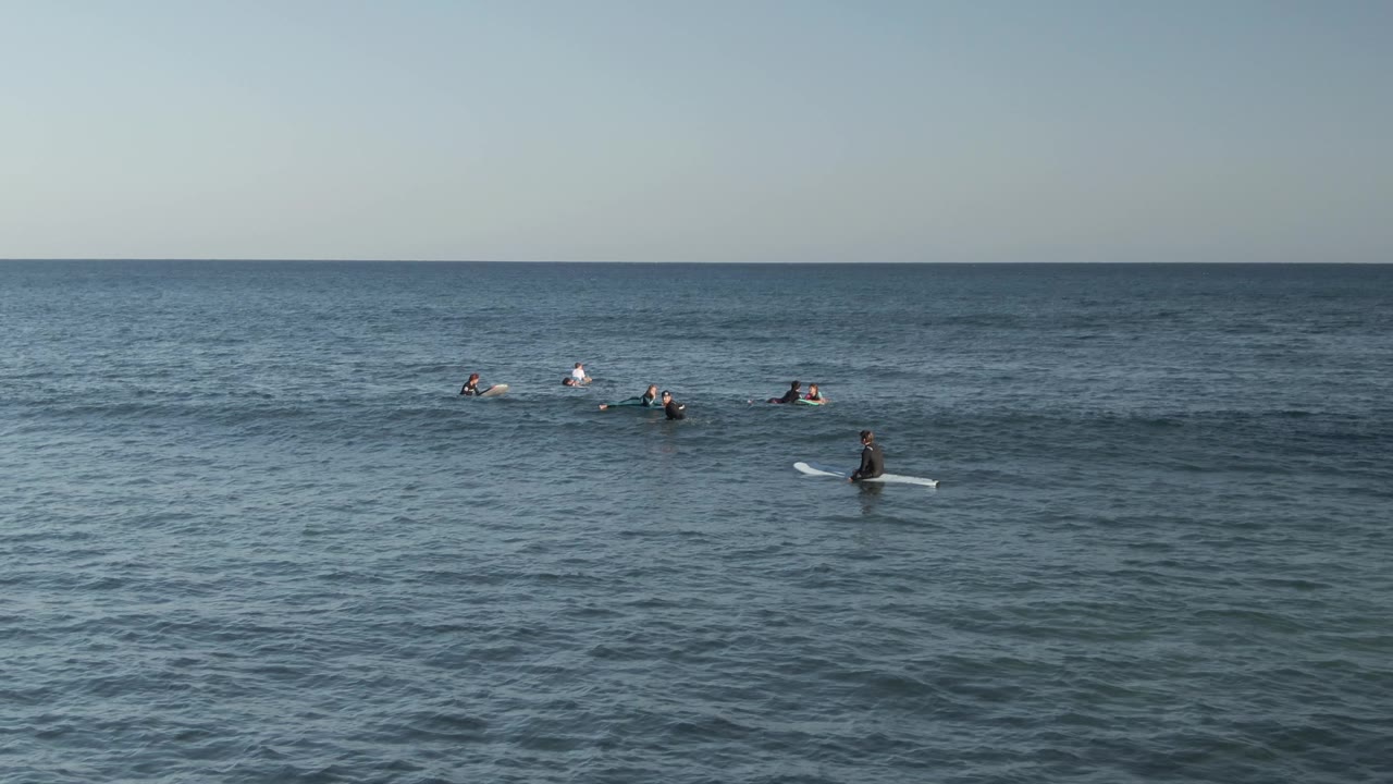 A wide Costa Azul Los Cabos ocean frame shows surfers on boards in small clusters, with darker rippled water behind the lineup and open sky above.