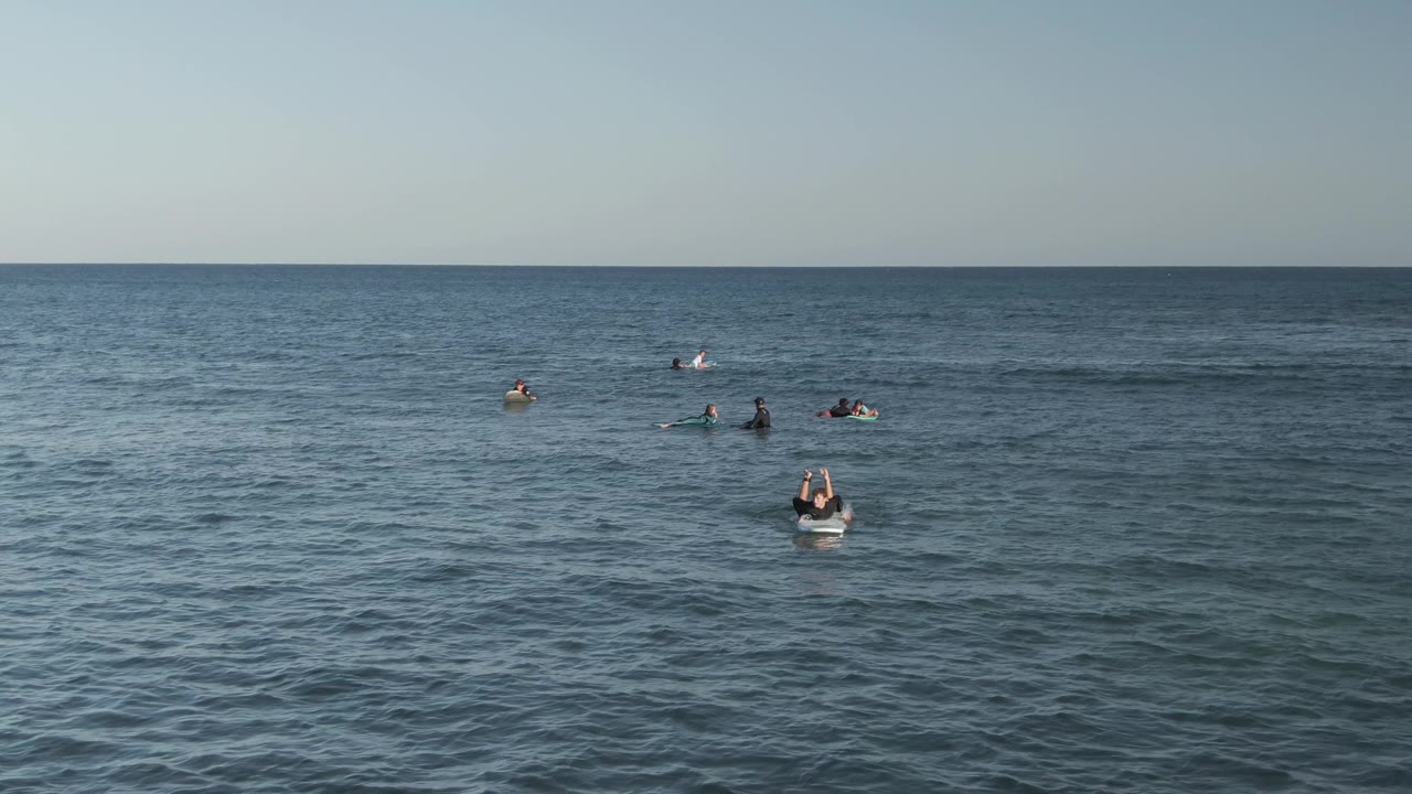 Wide elevated view of surfers on boards across calm ocean water at Costa Azul Los Cabos, with a pale sky and level horizon at the start of the clip.