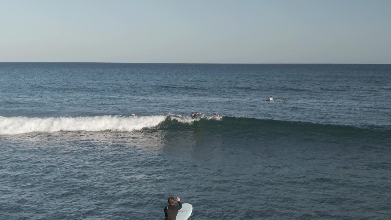 At Costa Azul Los Cabos, surfers sit and paddle beyond a white breaking wave while a foreground surfer in a dark wetsuit holds a white surfboard near the lower edge.