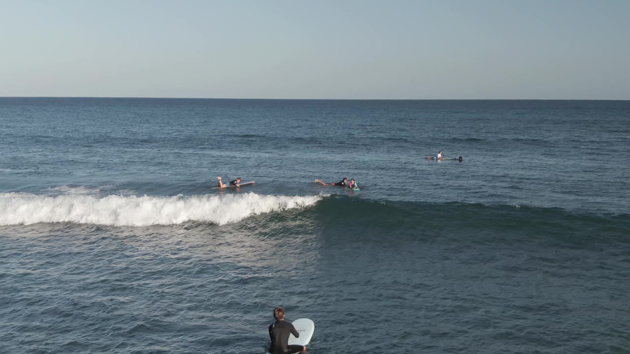 A wave breaks across the midground with surfers paddling and waiting offshore.