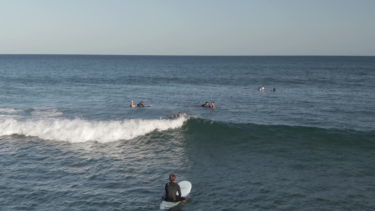 At Costa Azul Los Cabos, surfers are scattered beyond the break while a band of whitewater crosses the midground and the foreground surfer faces the open water.