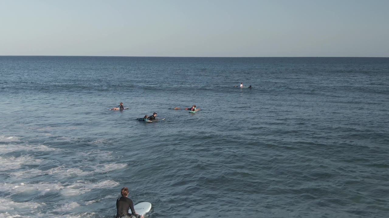 At Costa Azul Los Cabos, surfers sit offshore beyond low whitewater while a wetsuited person with a white surfboard stands near the lower foreground.