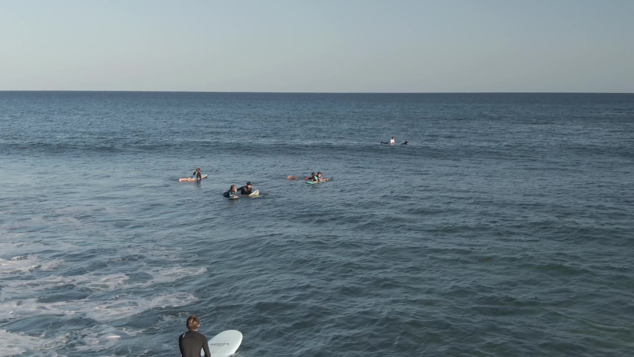 Several surfers on boards wait beyond small breaking waves.