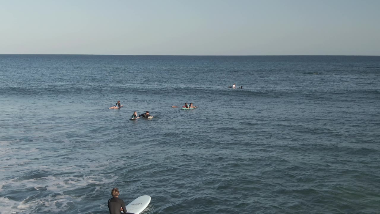 At Costa Azul Los Cabos, the ocean surface fills the frame with surfers waiting in small groups, a low wave line in the foreground, and a board-carrying person at the lower right.