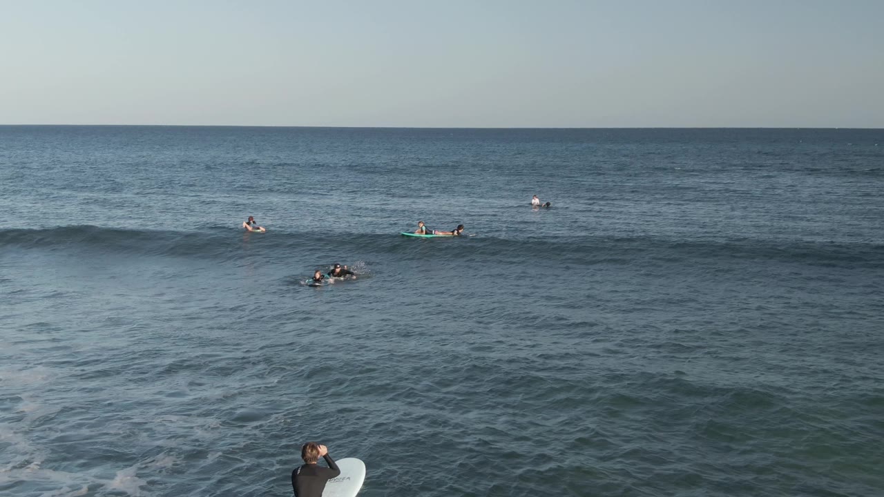 Wide ocean view at Costa Azul Los Cabos with several surfers on boards spread across the water and a small wave forming in the midground.