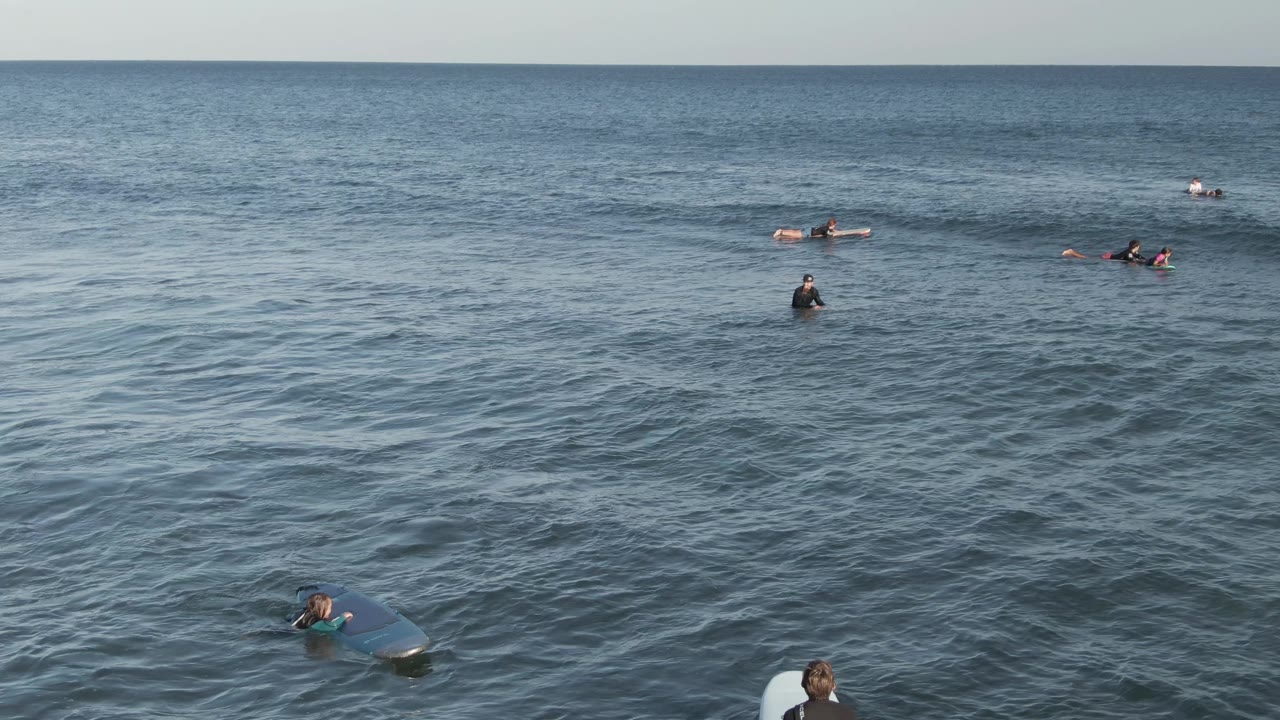 At Costa Azul Los Cabos, people on surfboards and bodyboards are visible across open ocean, with darker rippled water in the foreground and a clear horizon above.
