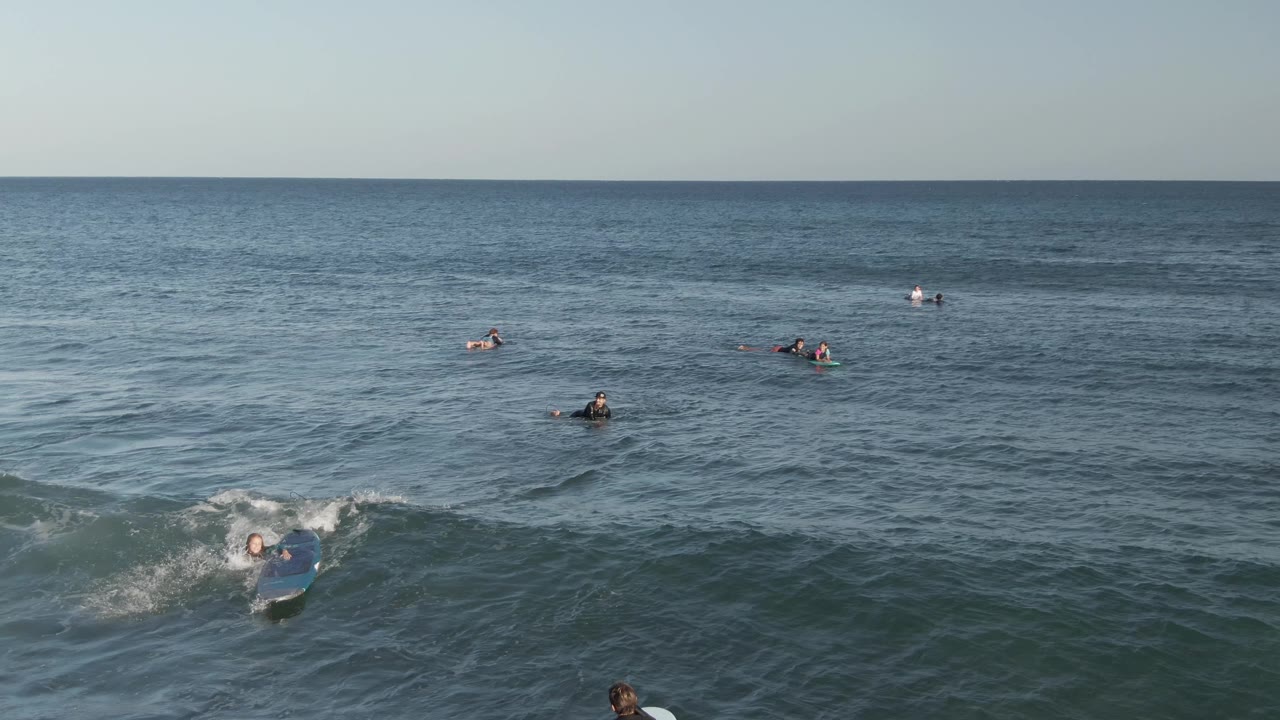 Wide elevated view of surfers in the ocean at Costa Azul Los Cabos, with a small wave breaking near the left edge and more boards scattered toward the horizon.