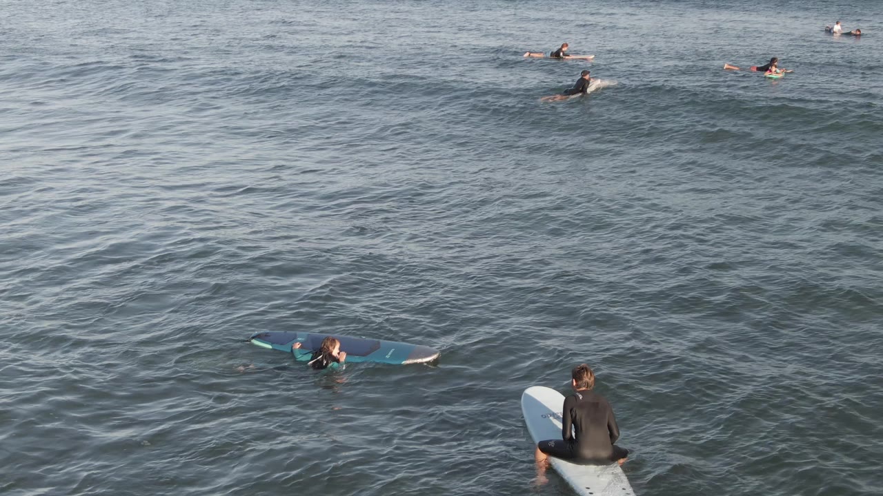 Surfers in wetsuits paddle and wait on surfboards in the ocean at Costa Azul Los Cabos, with one closer rider seated near the lower frame and several smaller surfers farther out.
