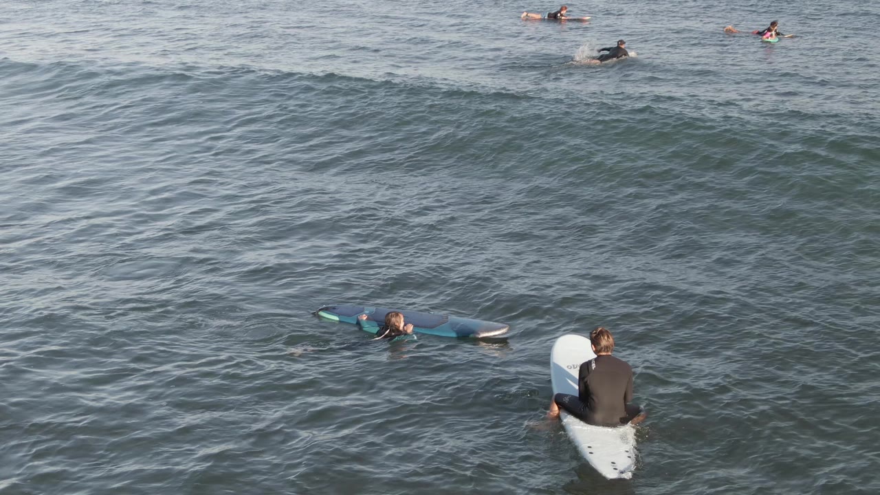 A close surfer sits facing away on a white surfboard while multiple distant surfers remain scattered along the upper part of the ocean at Costa Azul Los Cabos.