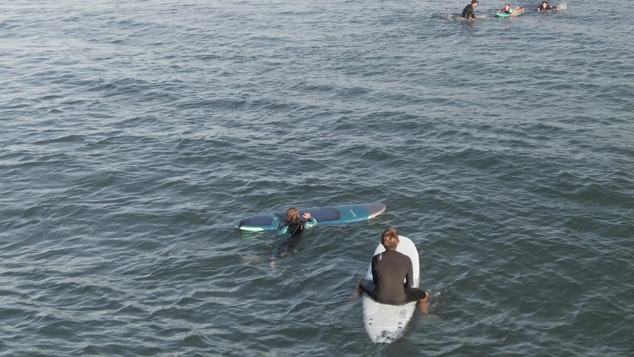 At Costa Azul Los Cabos, several wetsuited surfers float in blue-green ocean water, with one seated on a white board near the lower center and others farther out.