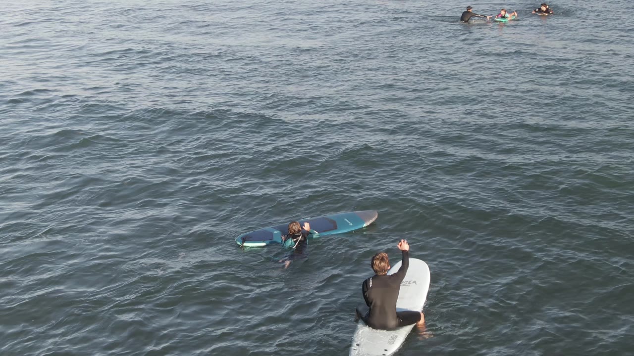 Wetsuited surfer raises an arm while another surfer waits on a blue board.
