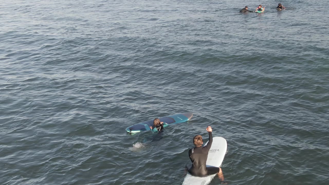 At Costa Azul Los Cabos, the raised-arm surfer remains near the lower right while the blue board sits to the left and small surfers are visible near the top edge.