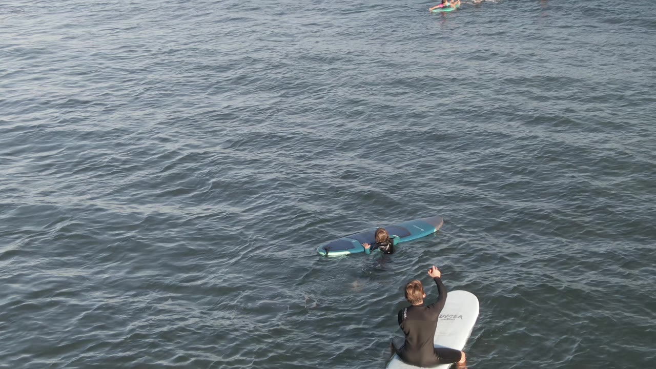 At Costa Azul Los Cabos, several surfers in wetsuits are visible in the ocean, including a foreground surfer sitting on a white board and another on a blue board farther out.