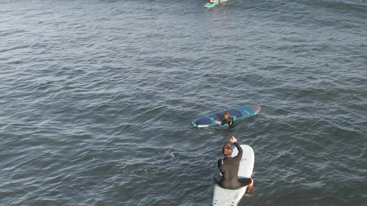At Costa Azul Los Cabos, the foreground surfer remains seated on a white surfboard while a darker swell line passes on the right and another surfer floats farther back.