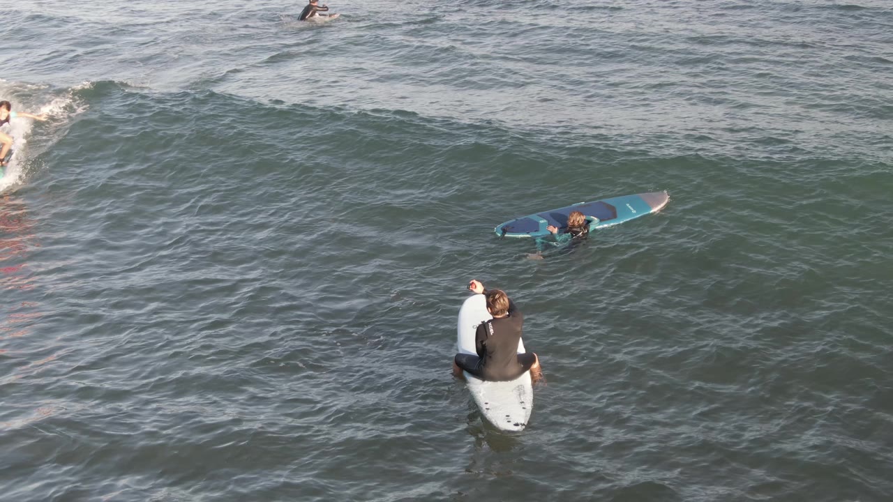 At Costa Azul Los Cabos, several surfers float in gray-green water while a nearer surfer sits on a white board in the lower part of the frame.