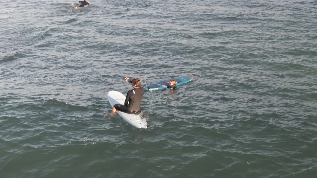 At Costa Azul Los Cabos, the nearest surfer sits on a white board while another surfer with a blue board floats nearby and small swells cross the frame.