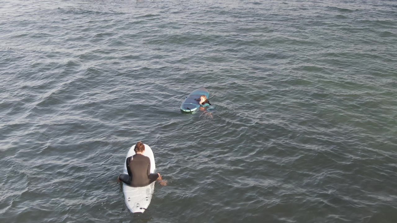 At Costa Azul Los Cabos, two surfers float in rippled green-gray water, with the white surfboard low in frame and the blue board farther out near the top.