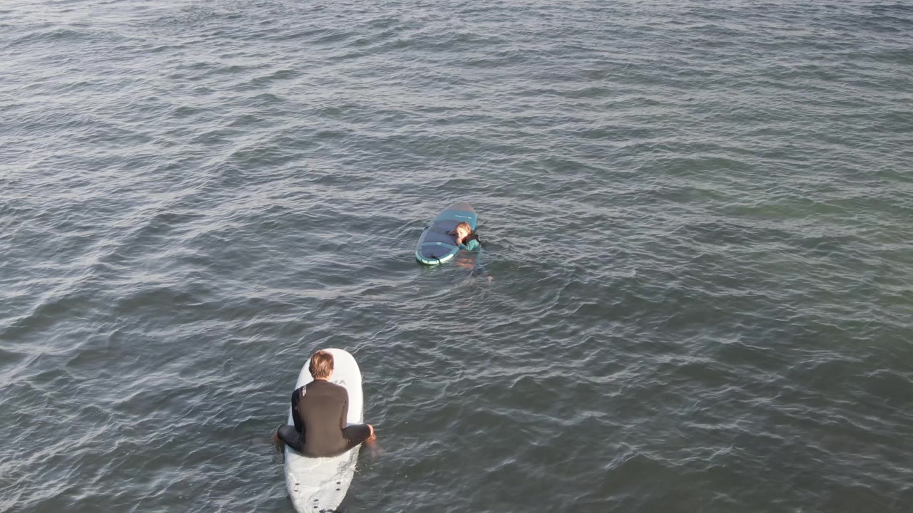 Foreground surfer seated on a white board with another surfer farther out.