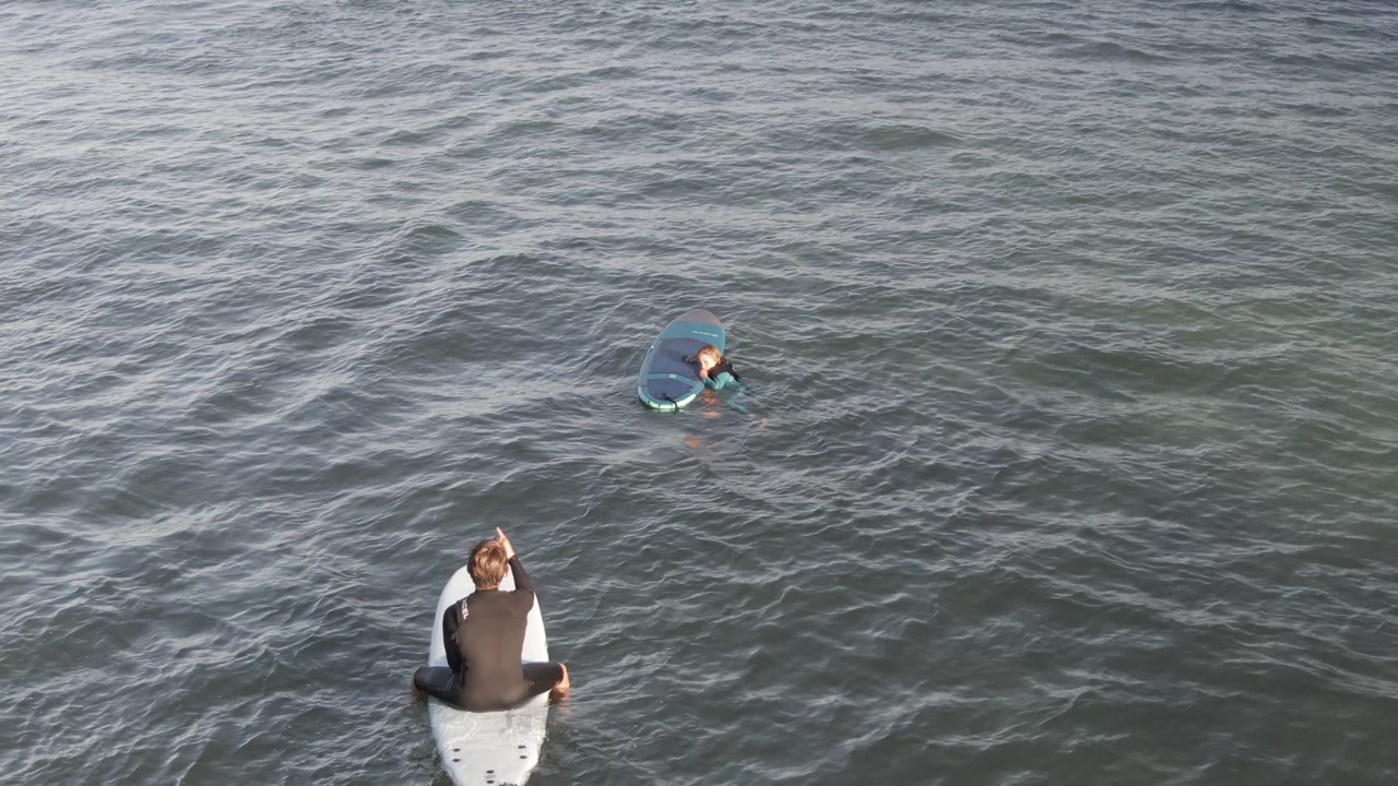 At Costa Azul Los Cabos, the foreground surfer on the white board raises an arm while the farther surfer stays near the upper frame on a blue board.