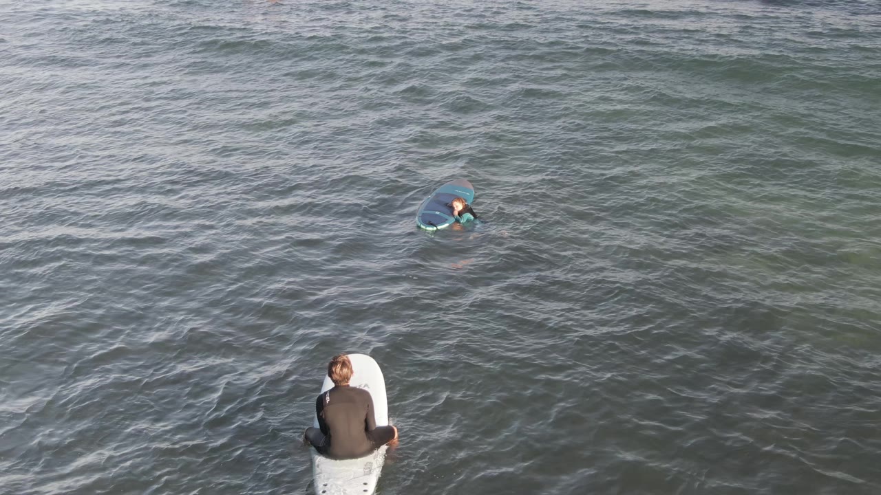 Aerial view of surfers in Costa Azul Los Cabos ocean water, with one surfer seated on a white board near the lower edge and another farther out paddling.