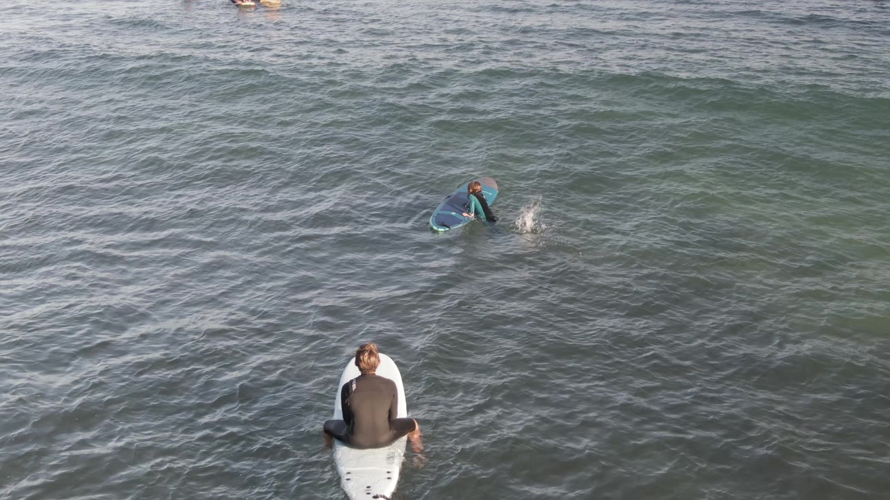 Surfer seated on a board while another paddles offshore.