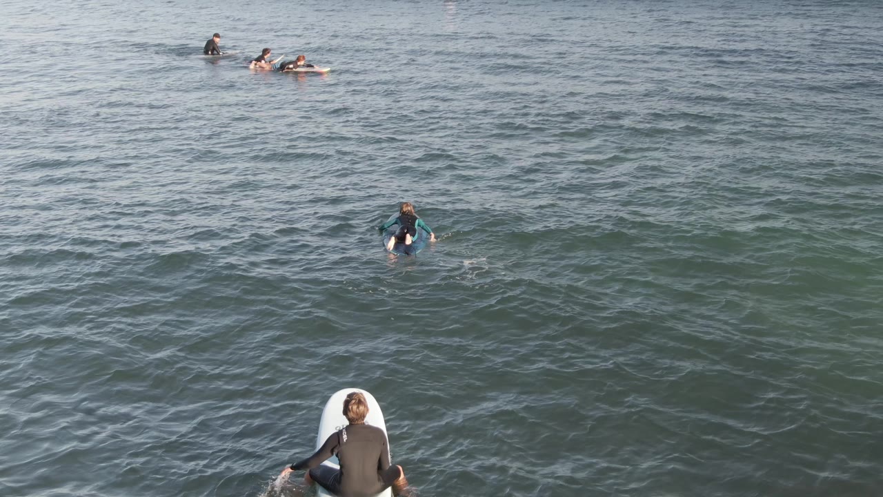 Surfers in wetsuits paddle and wait on surfboards in open water at Costa Azul Los Cabos, with a broad gray-blue ocean surface filling the frame.