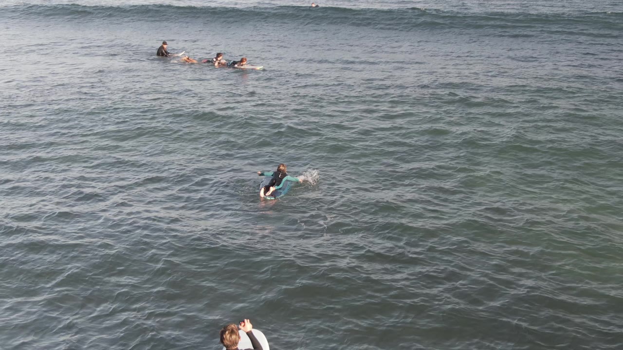 Multiple wetsuited surfers paddle across Costa Azul Los Cabos water as a small section of wave breaks in the background.