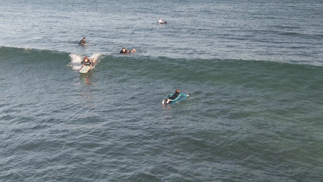 At Costa Azul Los Cabos, a surfer or bodyboarder rides near the top of a green wave as whitewater breaks behind them and other people float farther out.