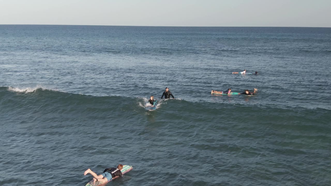 Surfers paddle and wait on surfboards in the ocean at Costa Azul Los Cabos, with a low wave cresting across the foreground and open water behind them.