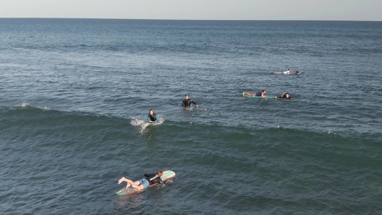 Multiple surfers remain spaced across the blue-green sea at Costa Azul Los Cabos, with one paddler closer to the lower frame and a gentle swell crossing the water.