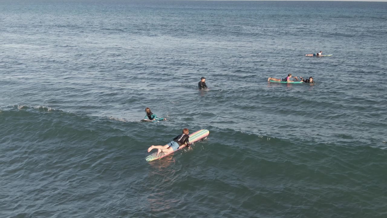 A foreground surfer paddles on a board at Costa Azul Los Cabos while several other surfers sit or paddle farther out beyond the small breaking wave.