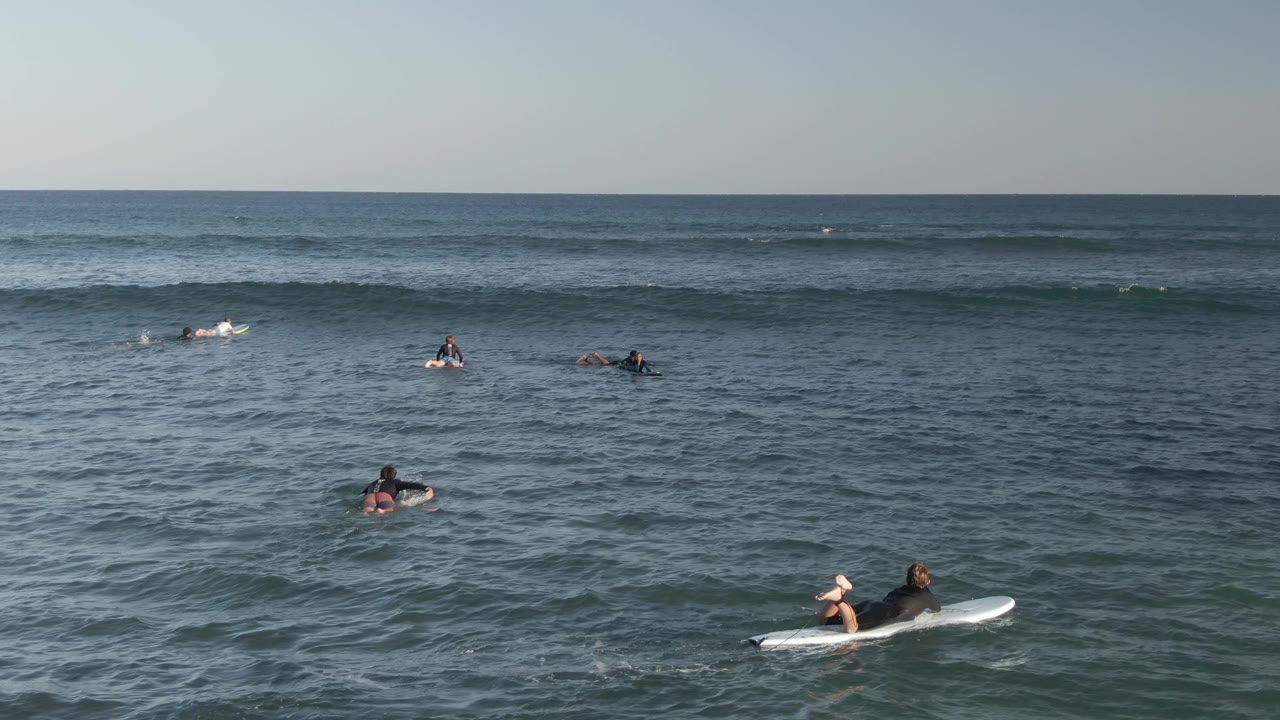 Wide elevated view of surfers at Costa Azul Los Cabos, Mexico, spread across blue-green ocean as a low swell line forms near the horizon.