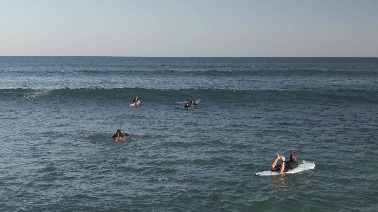 A foreground surfer lies on a white board at Costa Azul Los Cabos, Mexico, while other surfers wait farther out near small breaking waves.