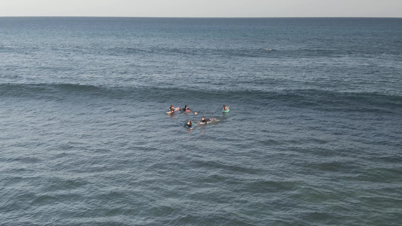 Wide aerial view at Costa Azul Los Cabos with small groups of surfers and bodyboarders floating in blue-green ocean water while a long swell line builds behind them.