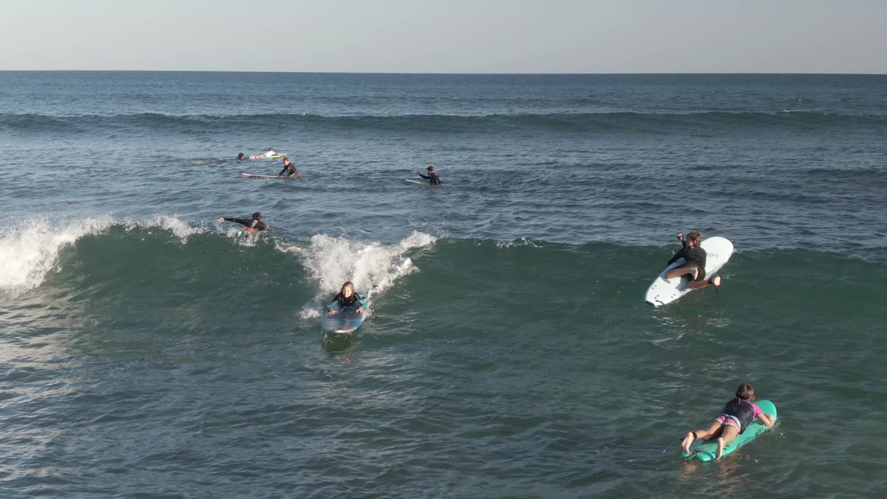 Aerial view at Costa Azul Los Cabos where a surfer on a white board rides near the right edge while other visible people paddle through the wave zone.