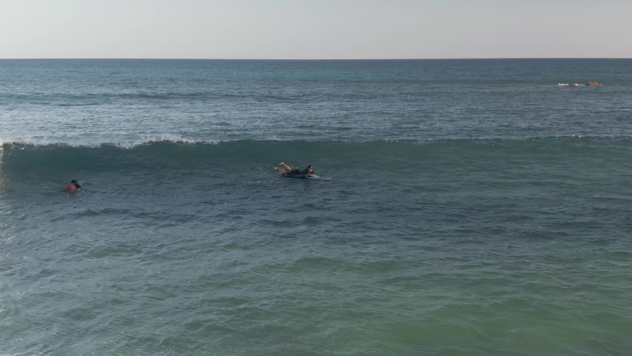 Distant surfers and bodyboarders wait and paddle in blue-green water at Costa Azul Los Cabos, with a low swell line forming across the frame.