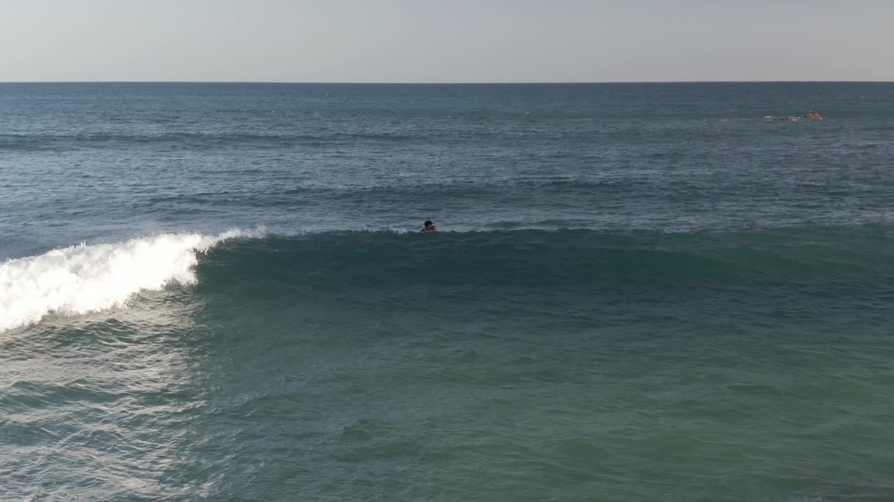 Whitewater breaks across the left side of a blue-green wave at Costa Azul Los Cabos, with a distant surfer visible beyond the foam line.