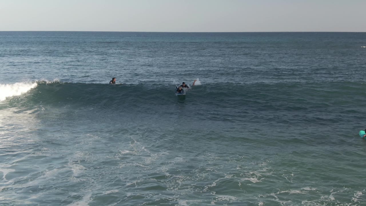 At Costa Azul Los Cabos, several surfers and bodyboarders sit or paddle near a green-blue wave beginning to break, with open ocean behind them.