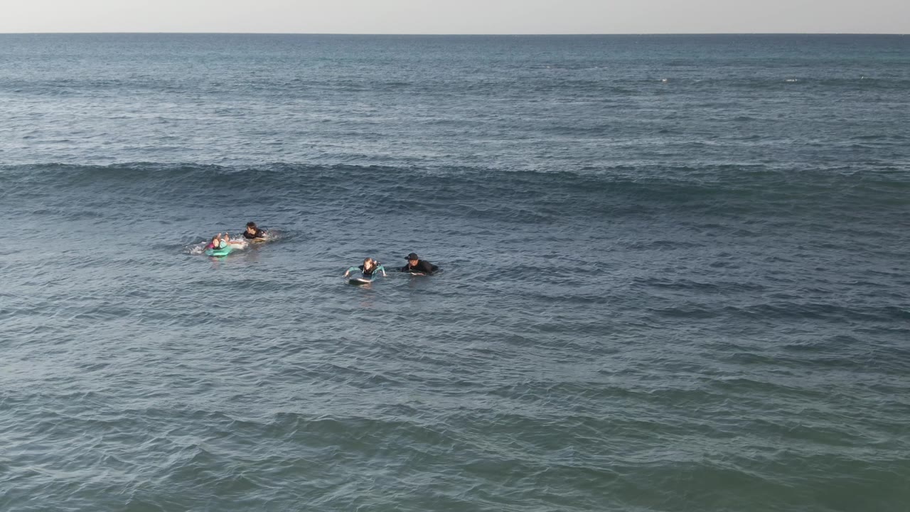At Costa Azul Los Cabos, several people paddle on boards in blue-green water as a dark swell forms across the frame.
