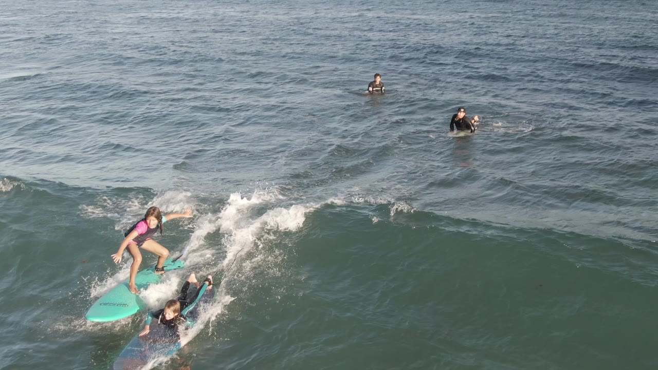At Costa Azul Los Cabos, a child rides a turquoise surfboard while an instructor paddles beside them in foamy green-blue water, with other surfers farther offshore.