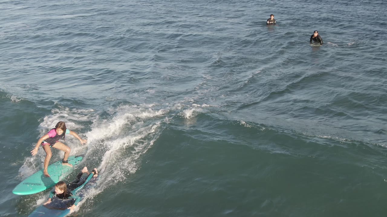 Child on a turquoise surfboard rides along a breaking wave near an instructor.